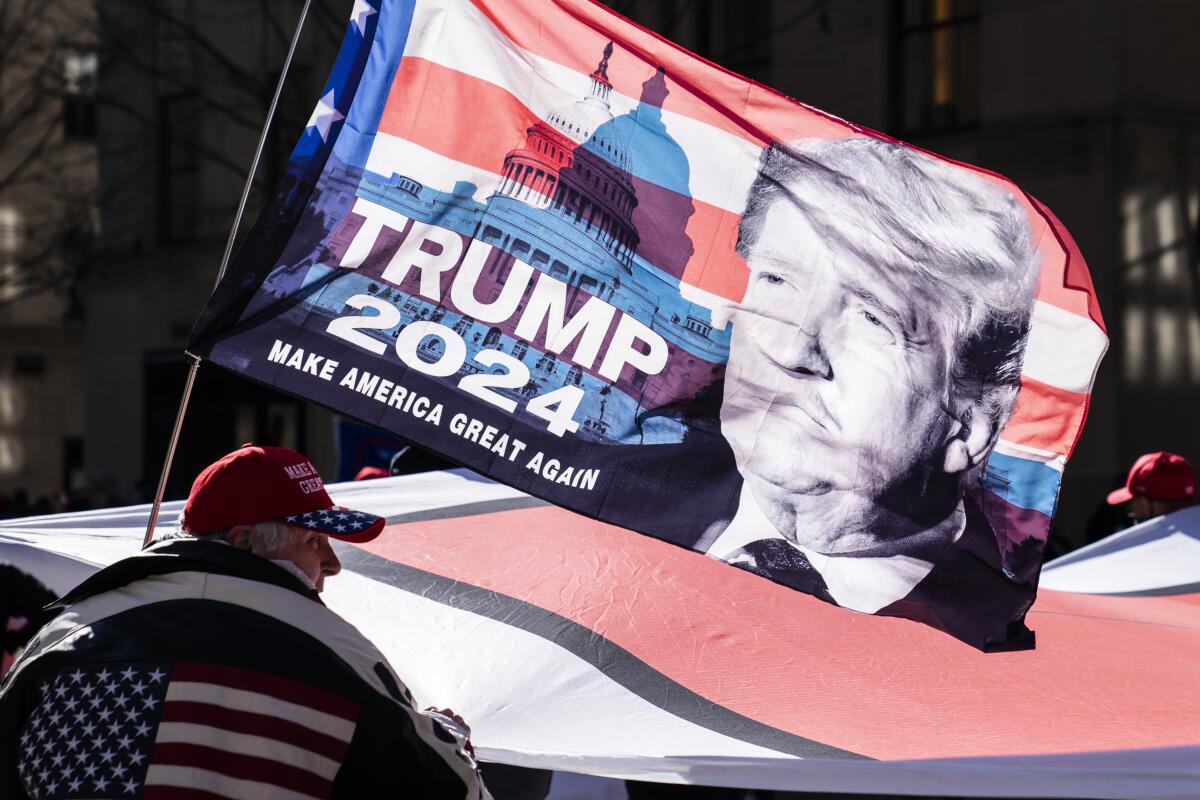 A man in a coat with a U.S. flag on int stands near a flag that says "Trump 2024: Make America Great Again."