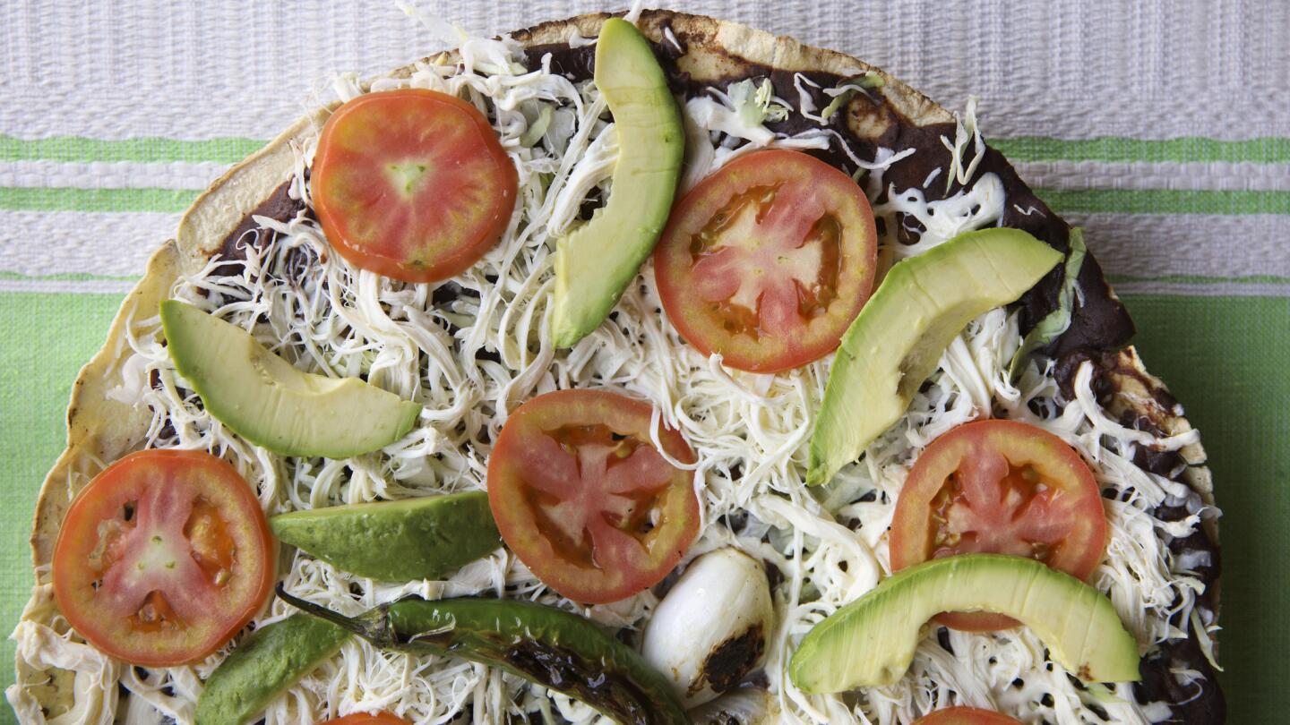 A vegetarian Tlayuda with avocado, tomato, beans, cabbage and cheese - topped with a fried serrano chile and a grilled Mexican green onion, is arranged for a photograph at Antequera de Oaxaca.