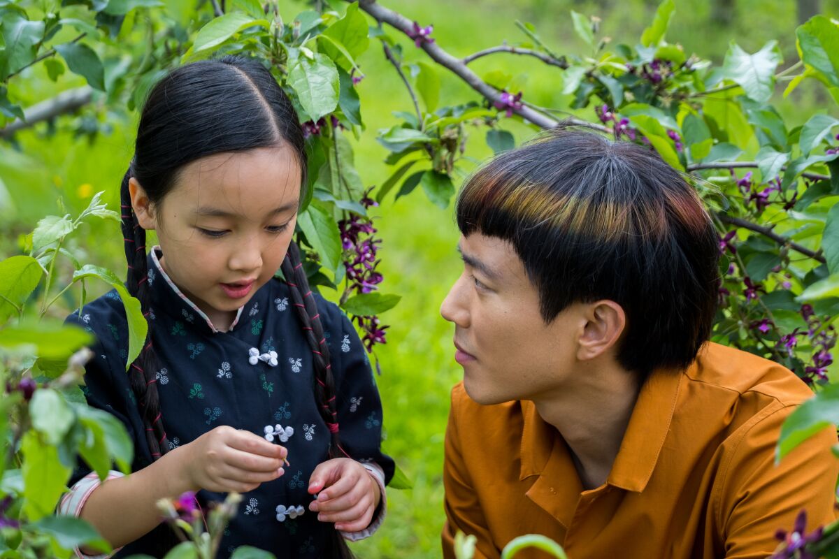 A girl plays with a twig under a branch while a man crouches beside her