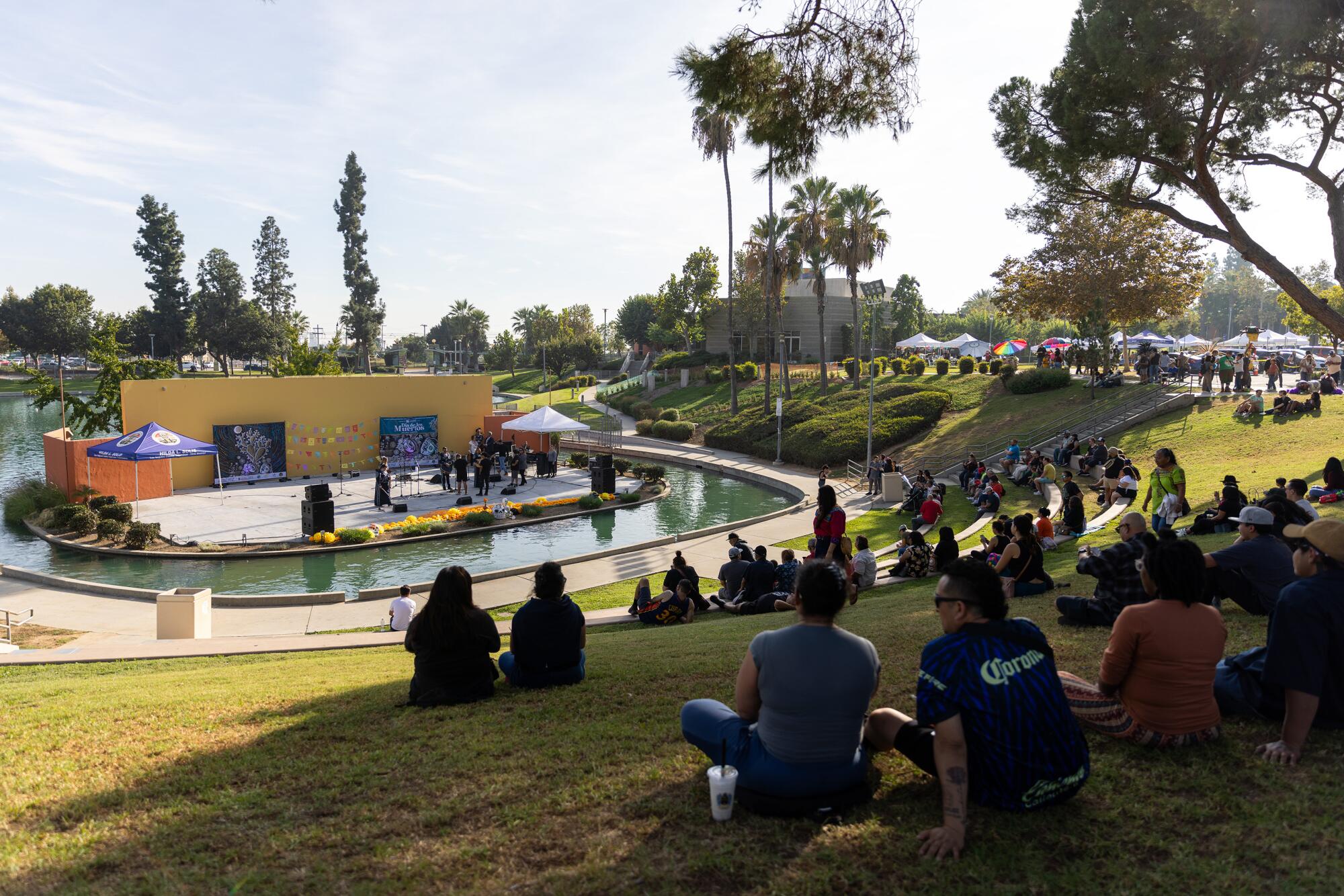 People sit on grassy hills around a stage, watching a performance in a park.