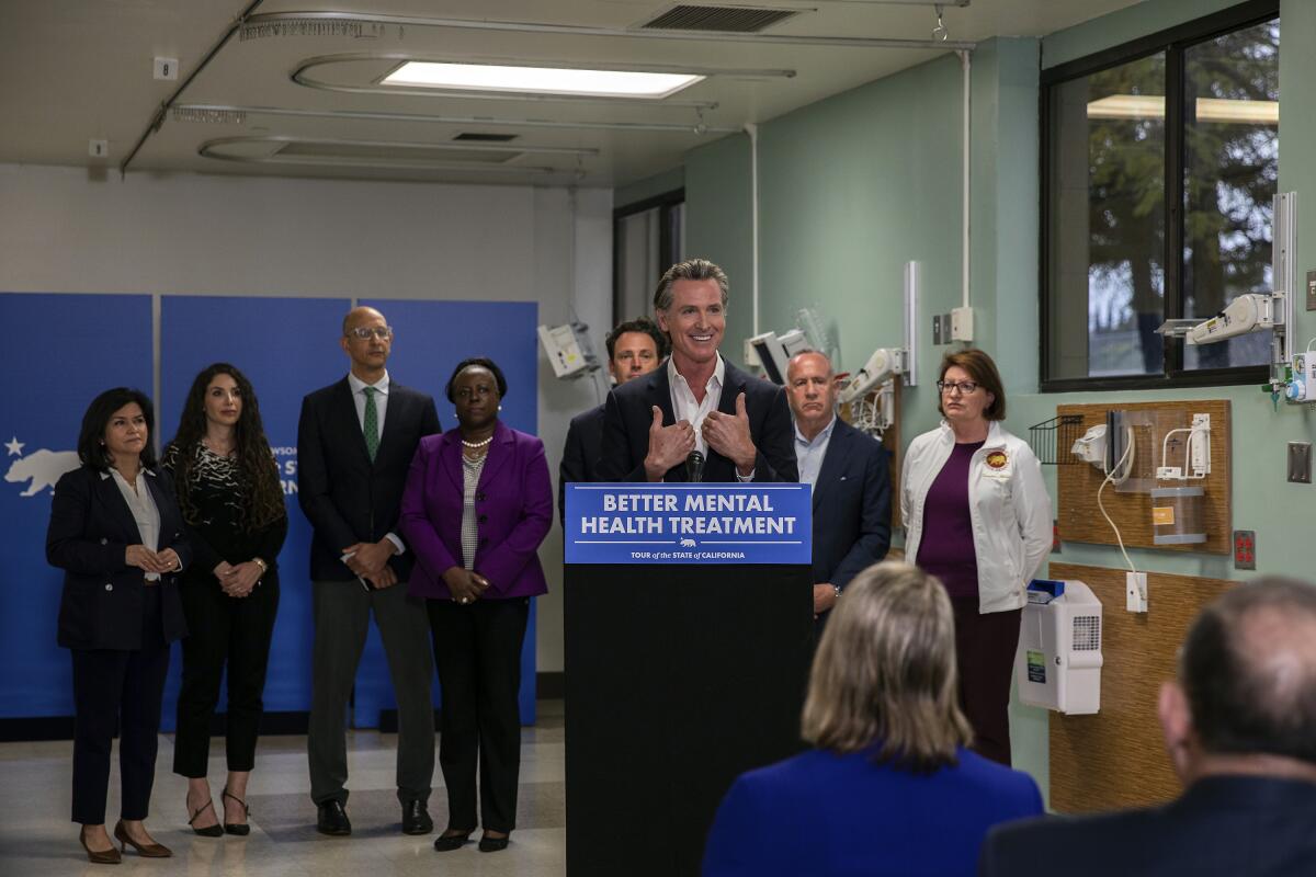 Gov. Gavin Newsom gestures as he speaks from a lectern with a sign reading "Better mental health treatment" as others look on
