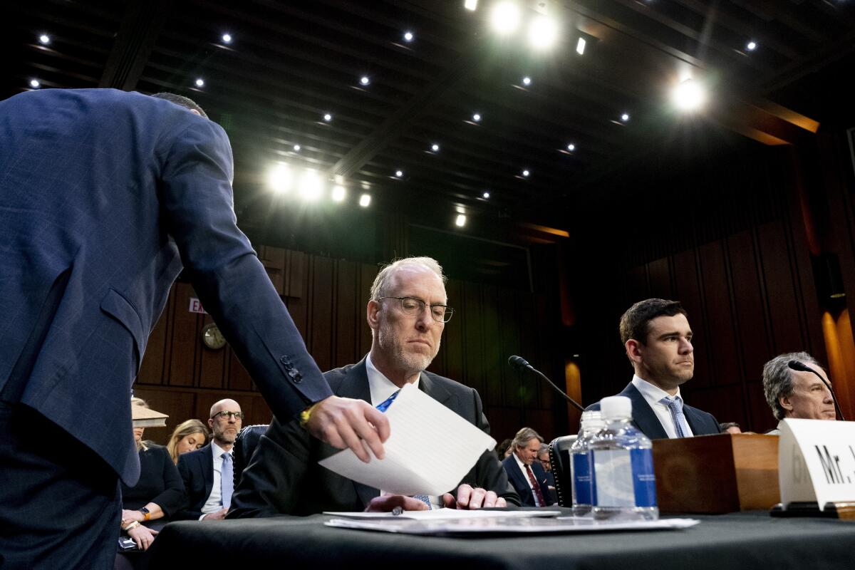 A man in a suit hands a document to another man seated at a desk inside a hearing room