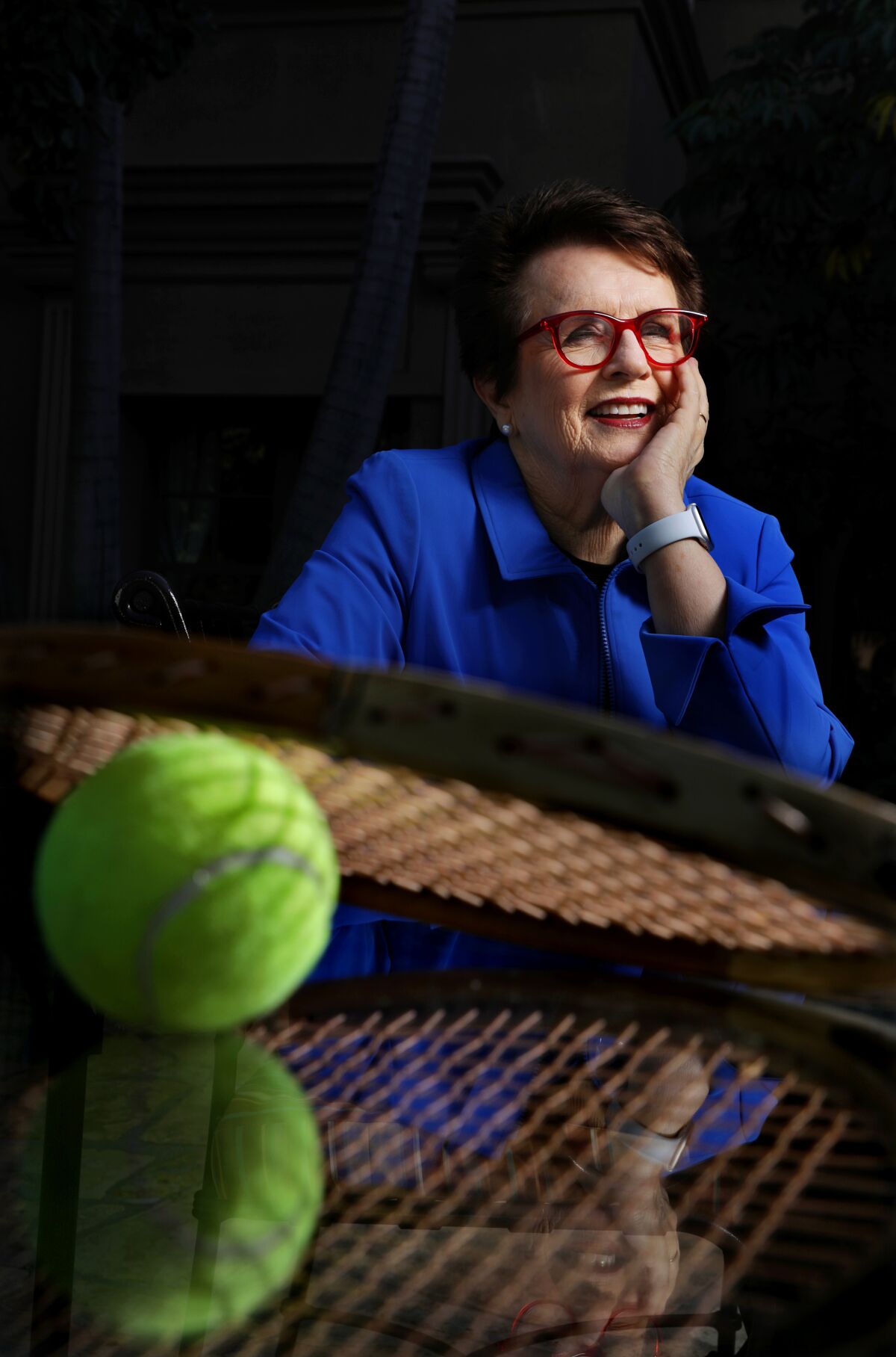 Billie Jean King in a blue jacket, seated, with a tennis ball and racket in the foreground.