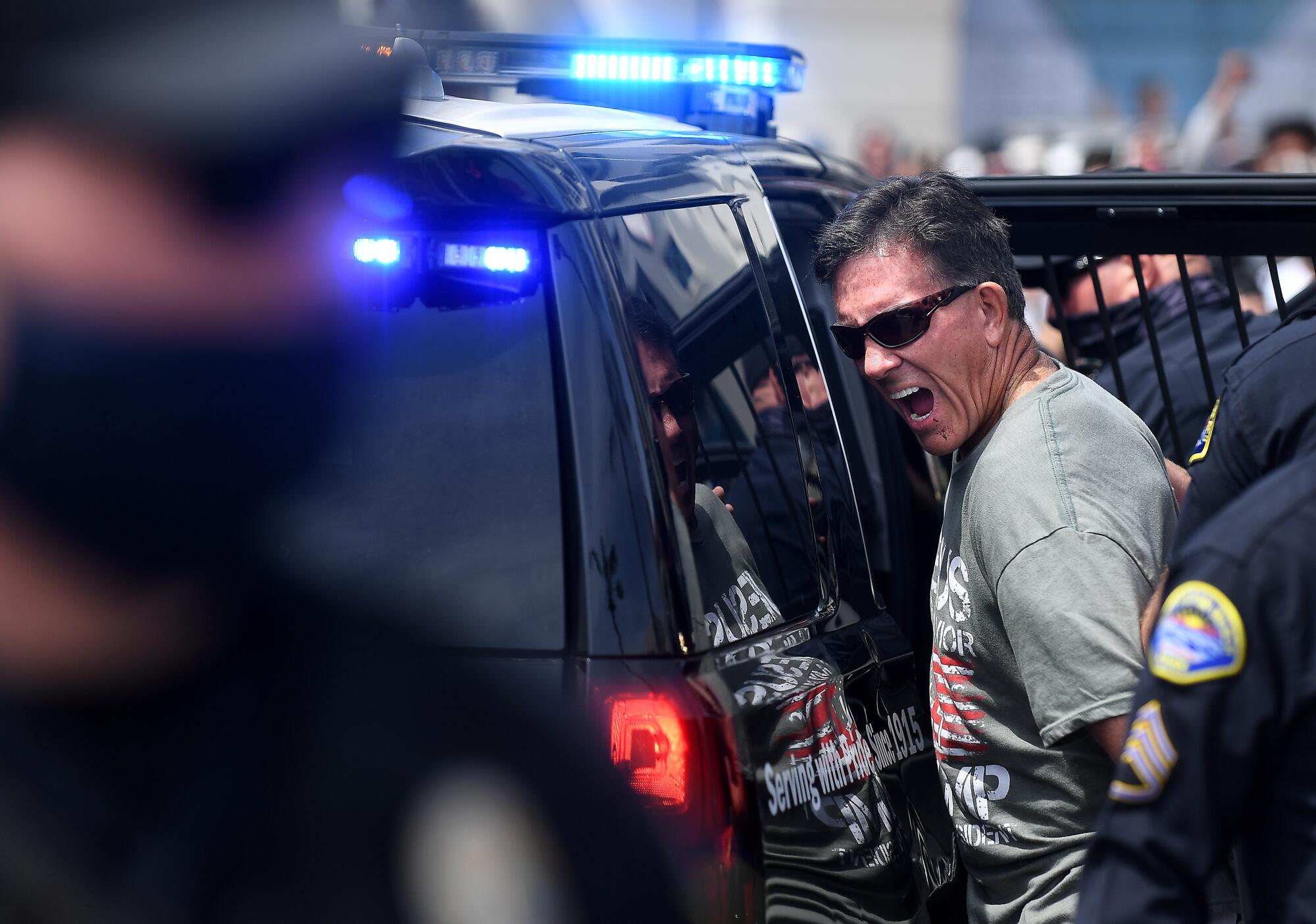 A man in sunglasses is ushered into the back of a police vehicle