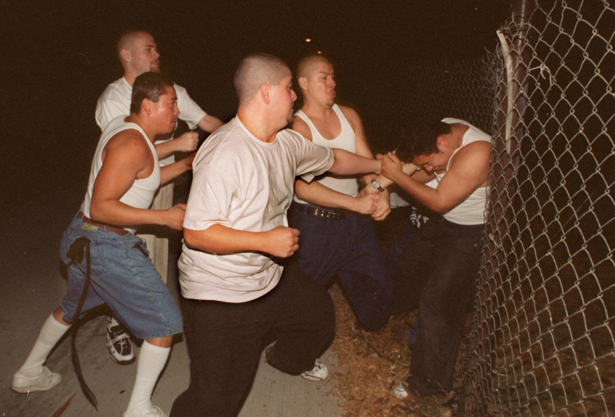 Four men in buzz cuts throw punches at a fifth, pinned against a chain link fence.