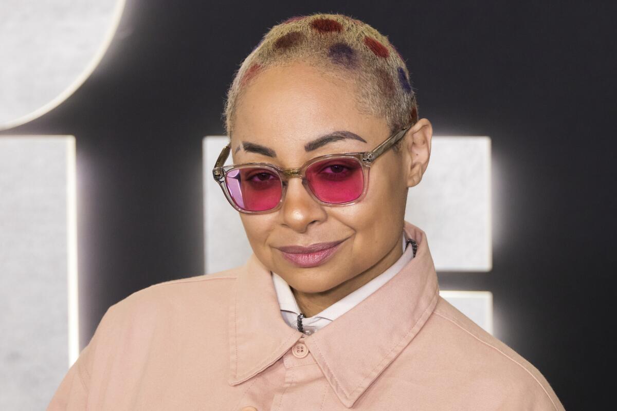Raven-Symoné smiles in pink sunglasses and a buttoned-up pink jacket with a patterned buzzcut.