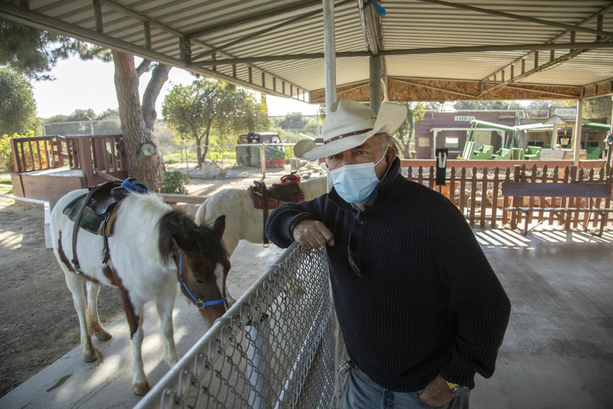 Rolando Wolovich in a Stetson hat and a mask leans on a fence next to two of his ponies