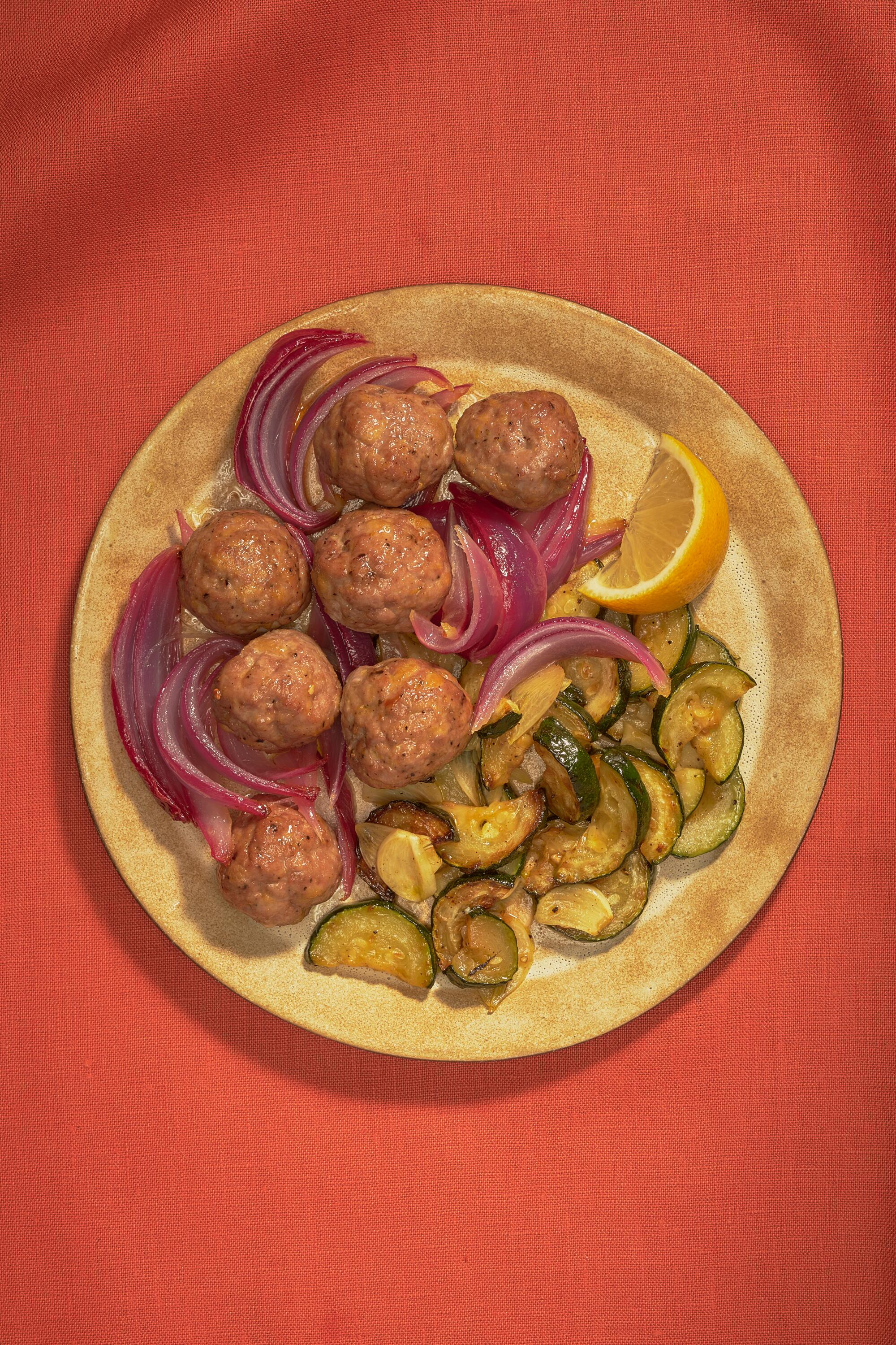 A plate of meatballs with red onions and zucchini on a red tablecloth