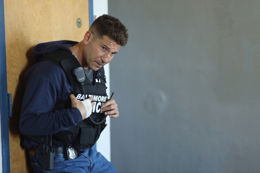 A man in a Baltimore Police vest leans against a door.