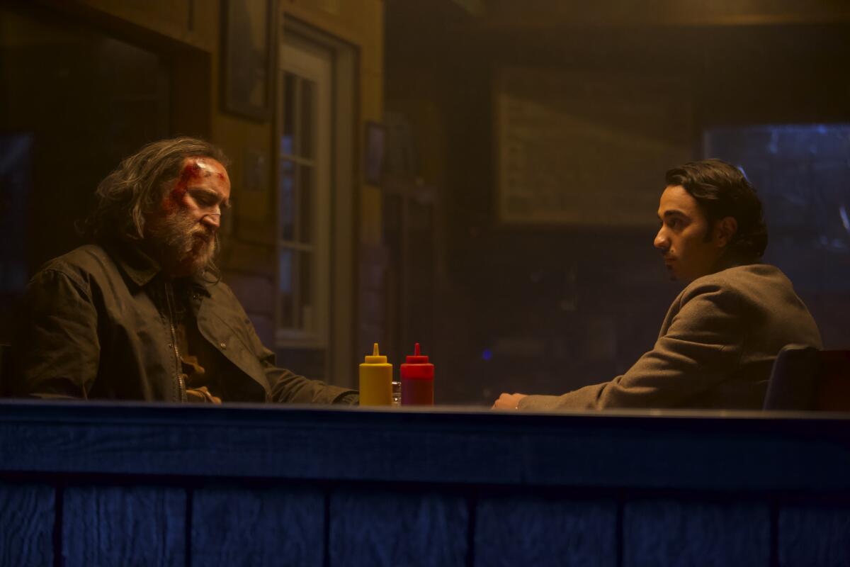 Seen through a window, a man, left, with blood on his face sits at a diner table with a man in a sport coat