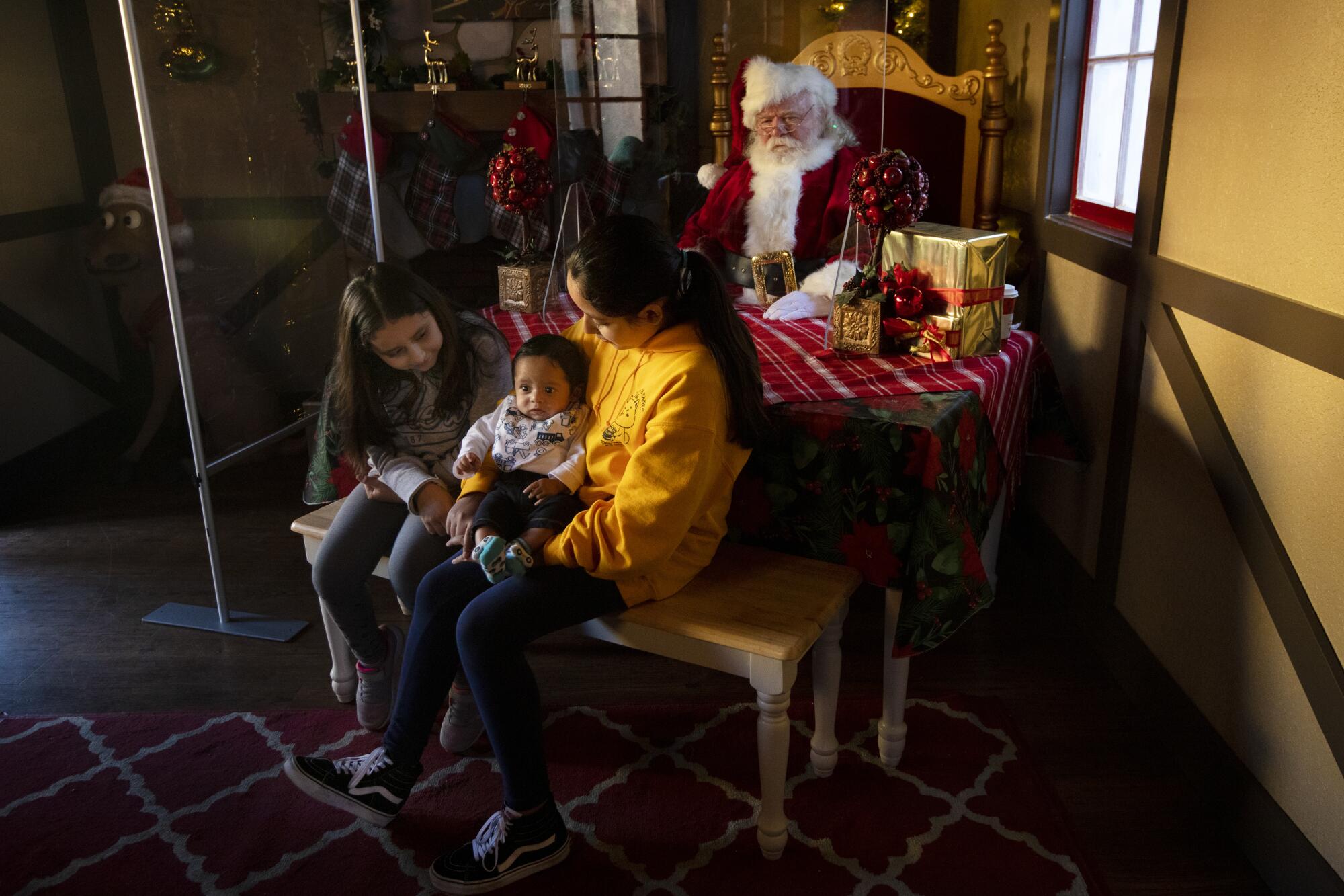 Two girls, one holding a smaller child, sit on a bench in front of a table, behind which Santa Claus sits.