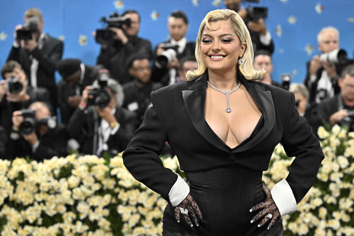Bebe Rexha in a black dress posing with her hands on her hips and smiling in front of a group of photographers