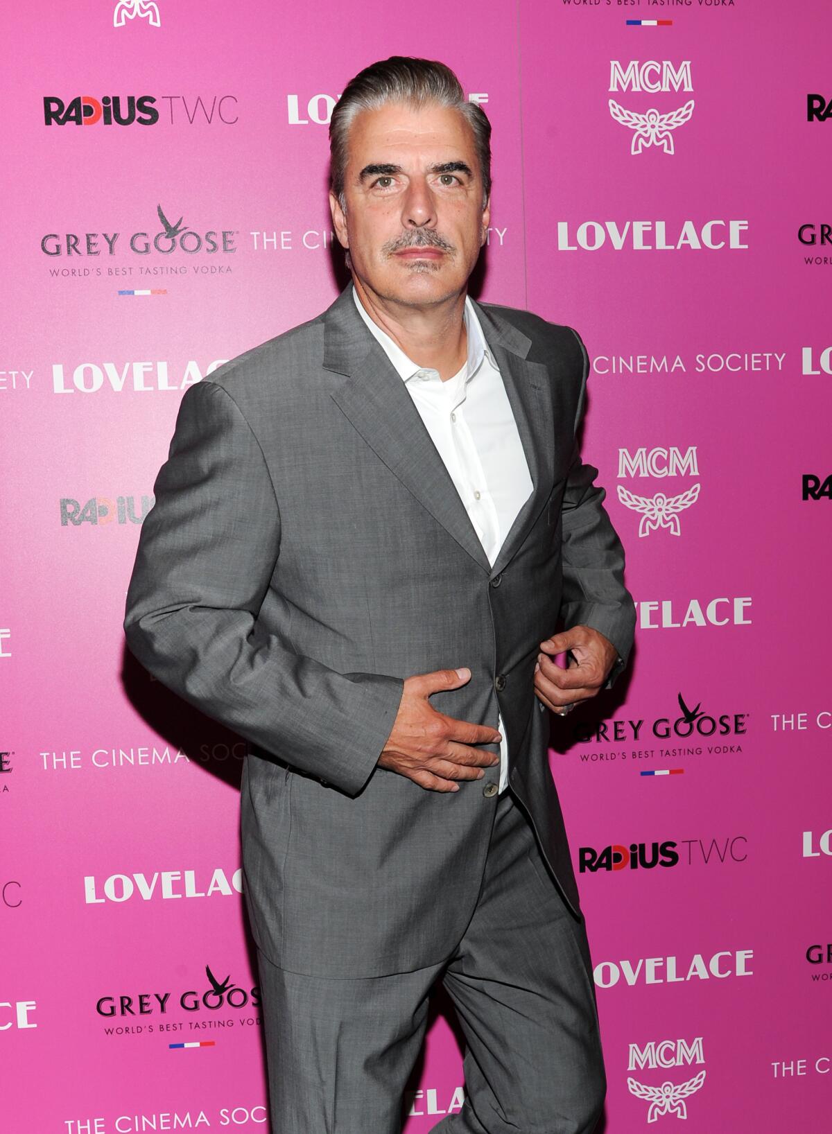 Chris Noth stands against a hot pink background in a gray suit and white shirt