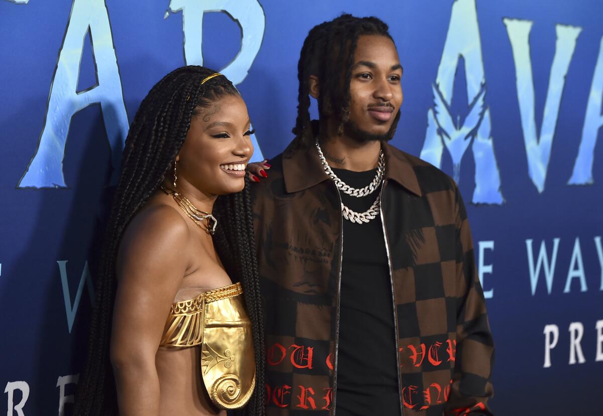 Halle Bailey in a gold top and stacked necklaces stands next to DDG in a brown jacket, black shirt and chains