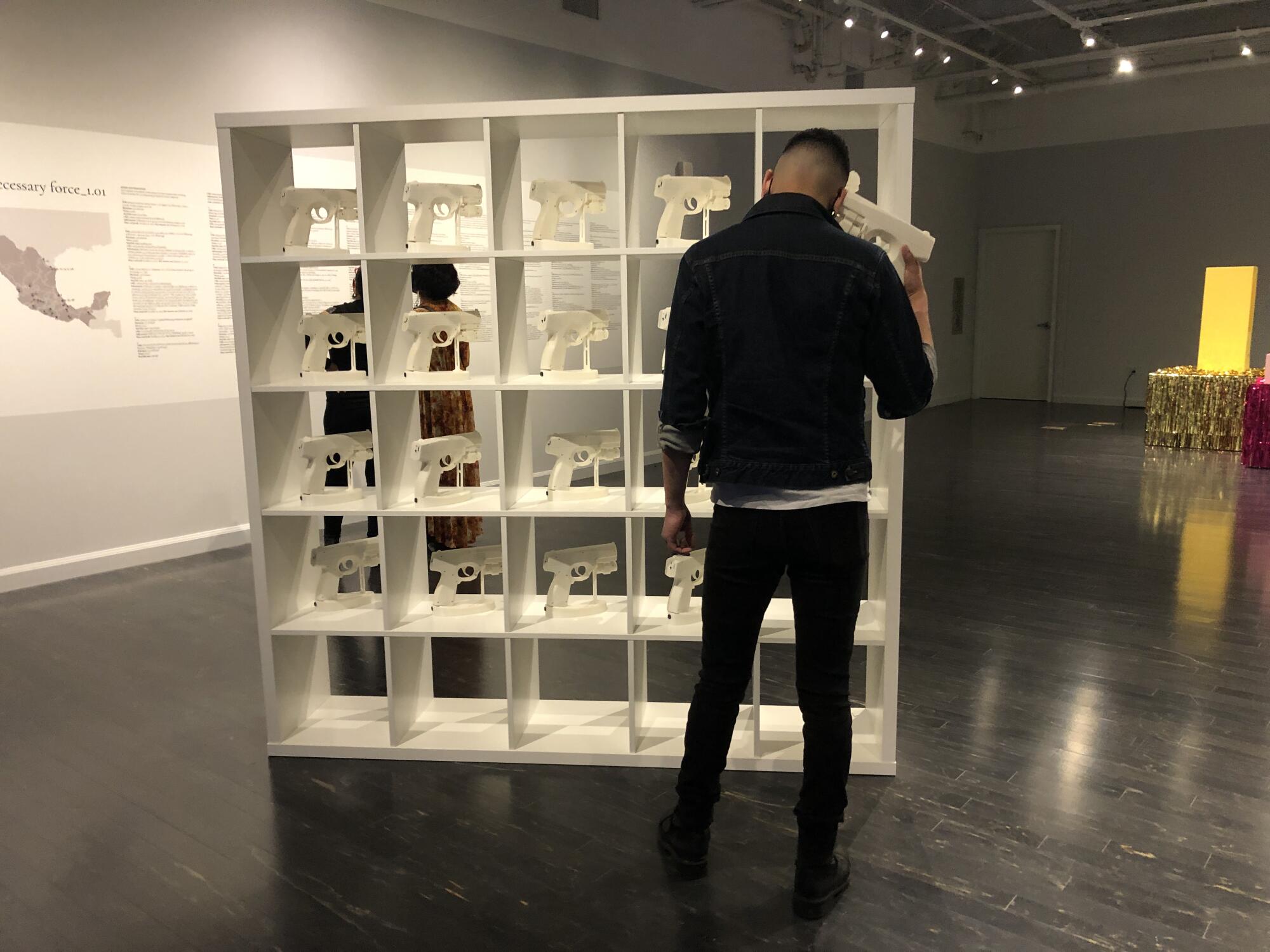 A museum visitor stands before a white cabinet bearing white gun-like objects, one of which he has pressed to his head