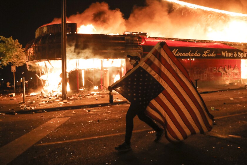 FILE - In this early Friday, May 29, 2020 file photo, a protester carries a U.S. flag upside, a sign of distress, next to a burning building in Minneapolis. Protests over the death of George Floyd, a black man who died in police custody Monday, broke out in Minneapolis and across the country. The U.S. has been dramatically disrupted in a matter of months, brought low by a global pandemic, Depression-era economic dislocation, and then, nationwide unrest over racial injustice. (AP Photo/Julio Cortez)
