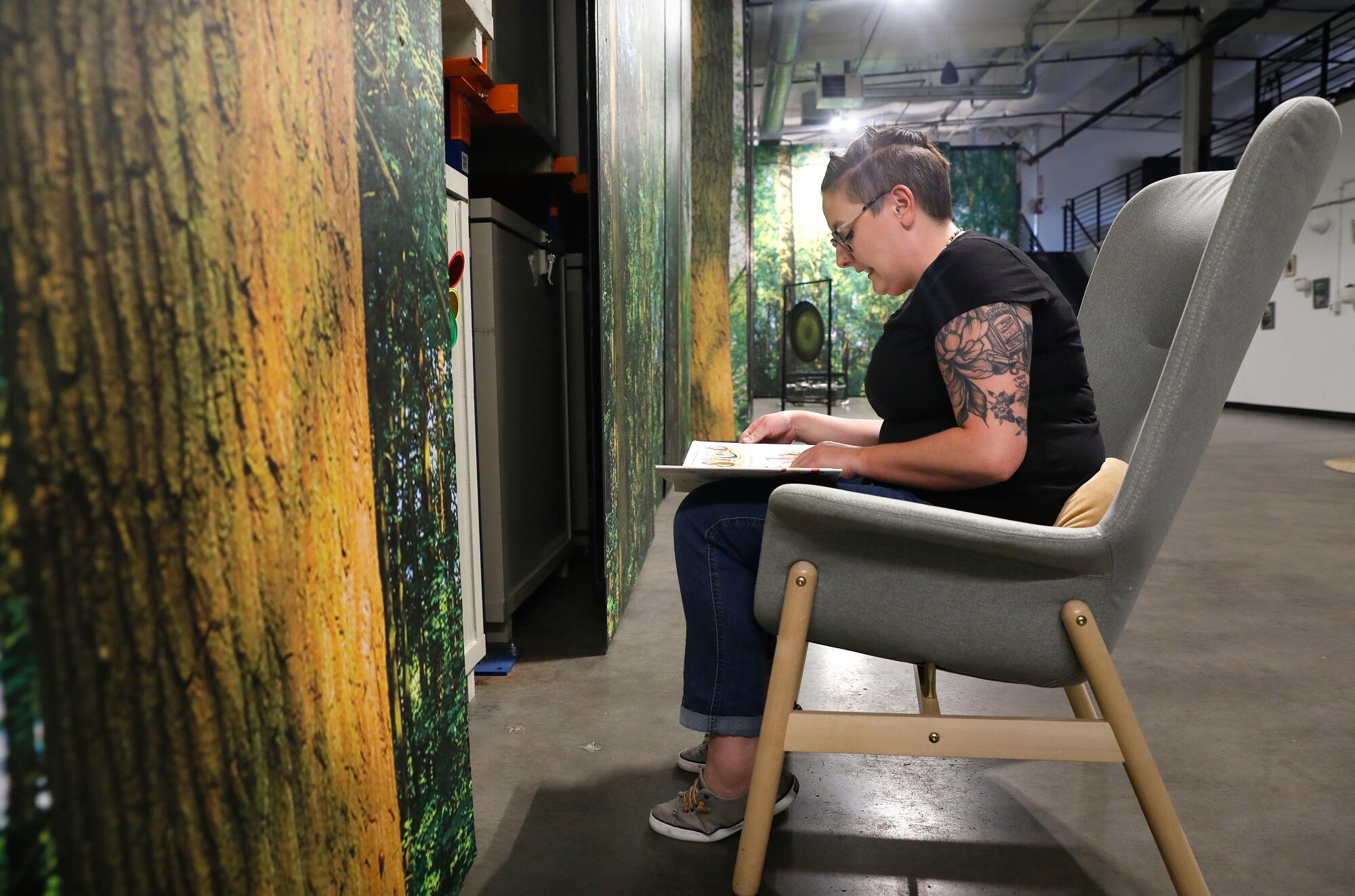 Katey Houston sits in a chair reading a book to children undergoing human composting in front of forest-themed panels.