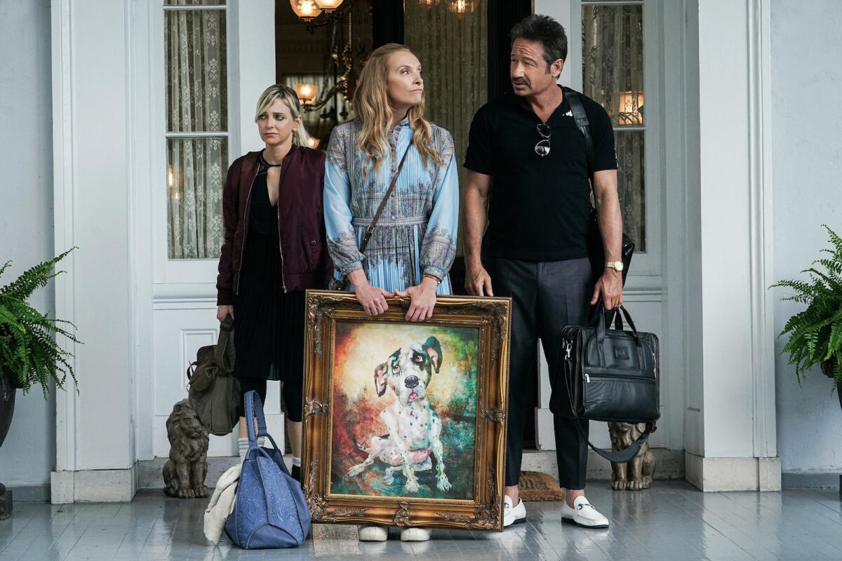 Two women and a man stand at the entrance to a large house holding their belongings.