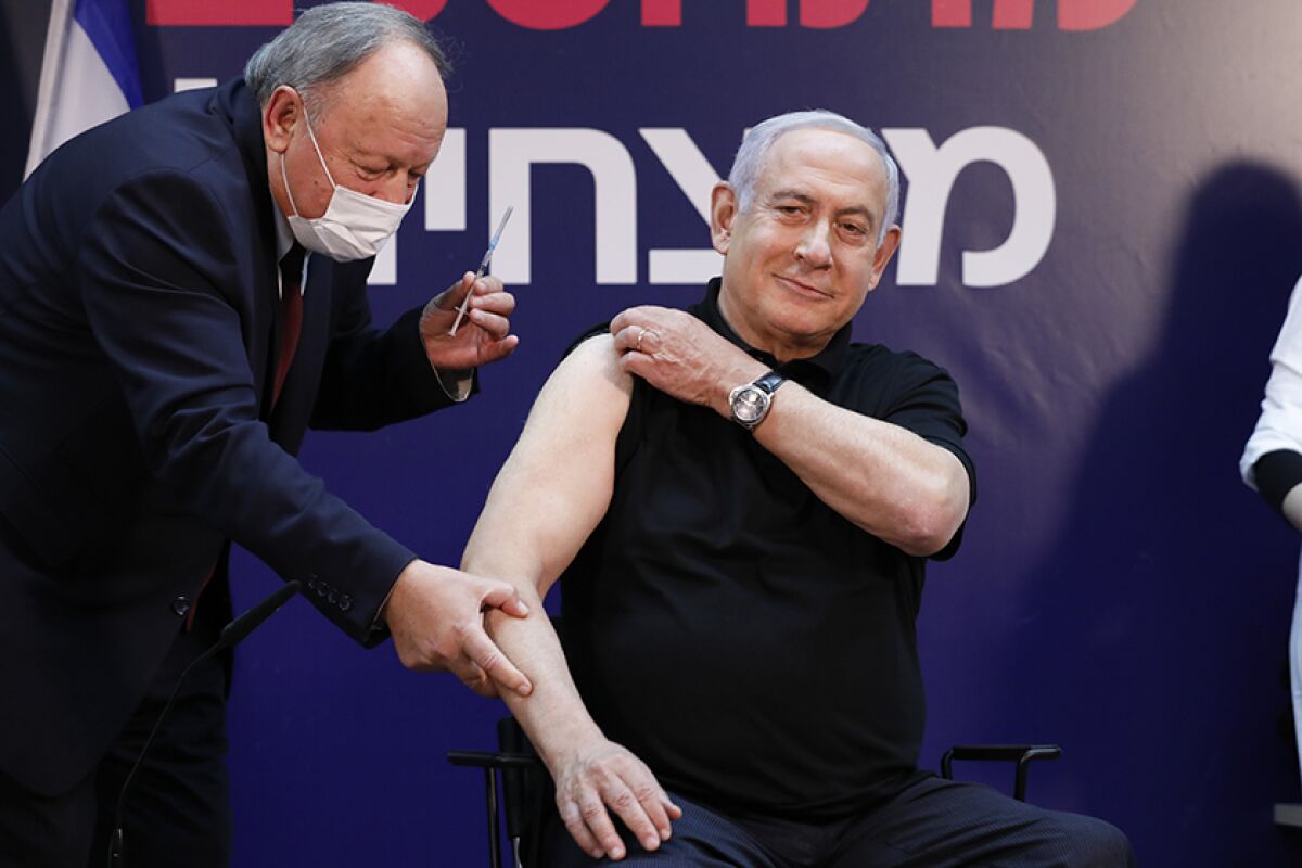 Benjamin Netanyahu rolls up his sleeve and smiles as a man in a mask holds a vaccine shot.