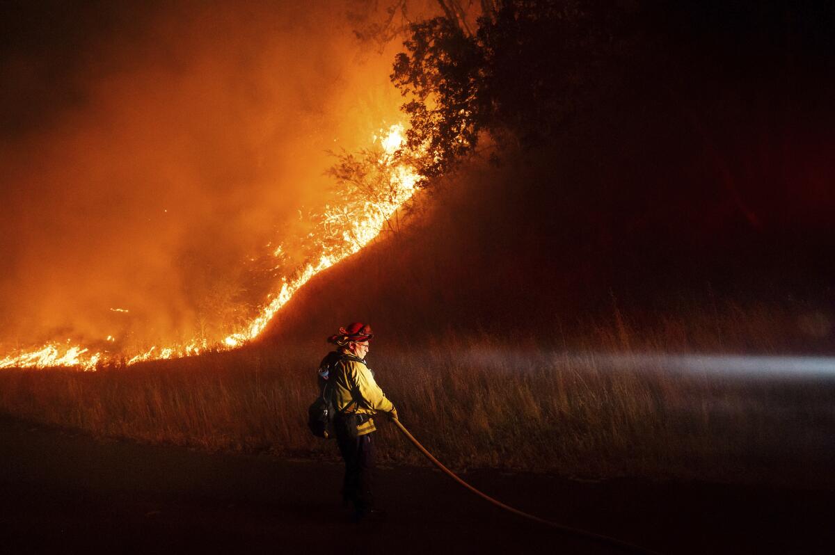 A firefighter backdropped by a line of flames holds a hose.
