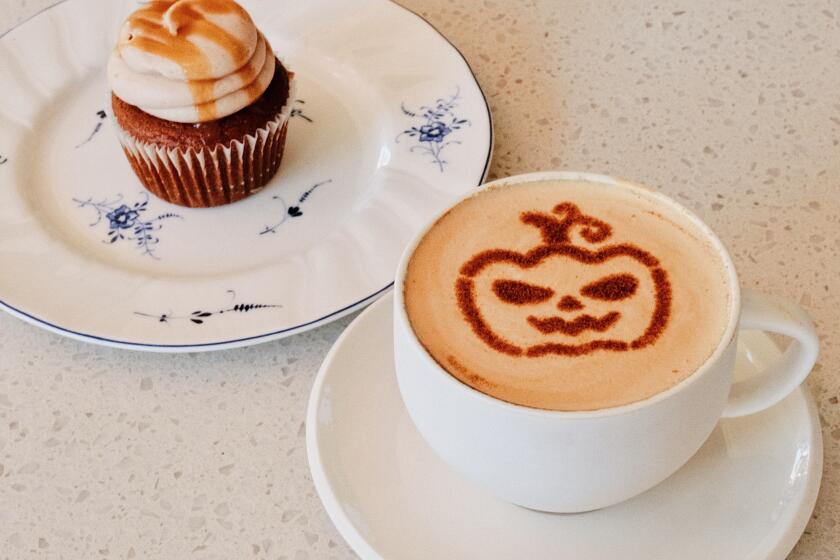 A latte in a white mug topped with a cinnamon stencil of a pumpkin at Nonna Mercato in Long Beach. Behind, a pumpkin cupcake