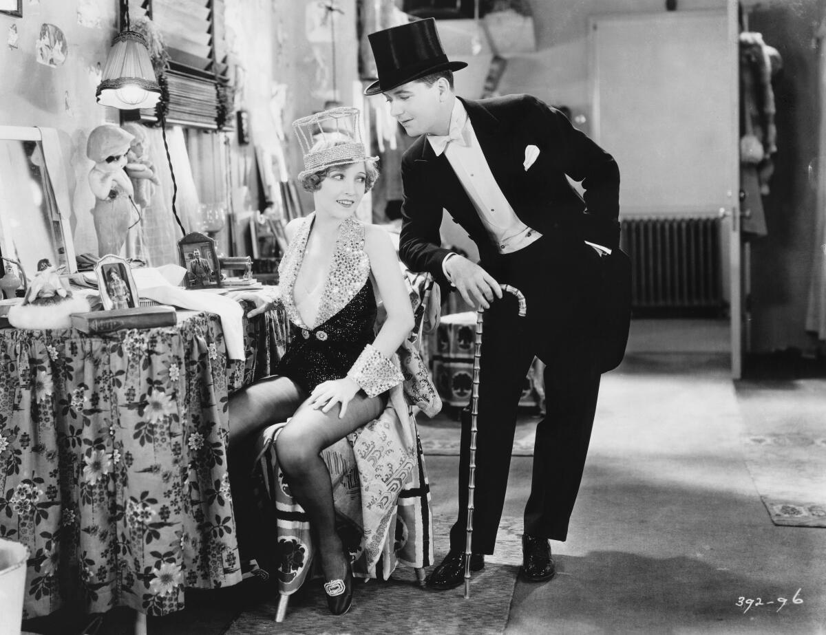 A man in top hat and tails leans over a woman seated at a dressing table backstage.