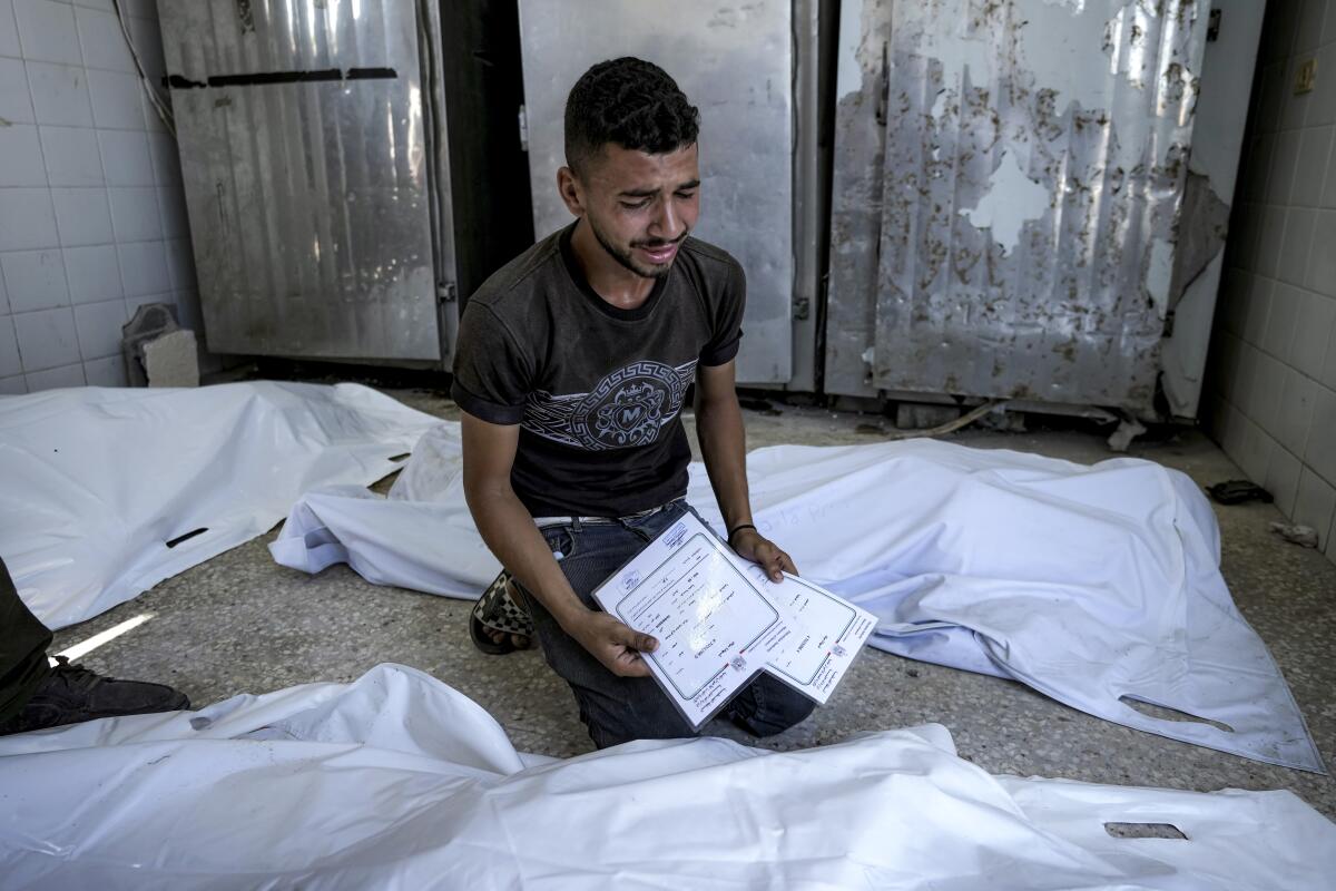 A Palestinian man sits by bodies covered in white sheets in a morgue. 