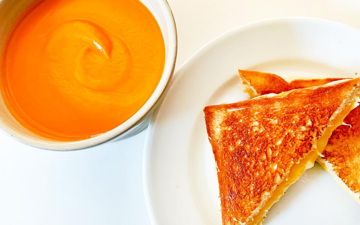 A bowl of creamy tomato soup beside a plate with a grilled-cheese sandwich.