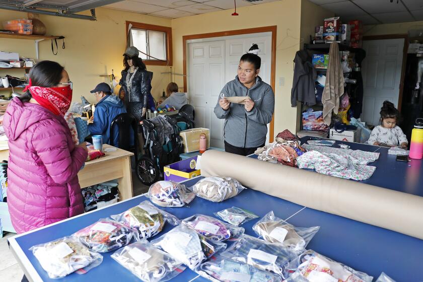 Trisha Garay, center, talks to Amy Guei, left, a speech therapist at Hoag Hospital, while she picks up face masks as well as donate some linen, at Garay's home in Fountain Valley on Friday, April 10. Garay has been making hundreds of homemade masks a day, out of her garage, to give away for free to her community. Medical offices, postal workers, bus drivers and pharmacists have all "placed orders" with her. She makes the masks on a factory sewing machine at home with her brother Danny Nguyen, background left, and his wife Kristine Tran, background right. At far right is Garay's 2-year-old daughter Adele.