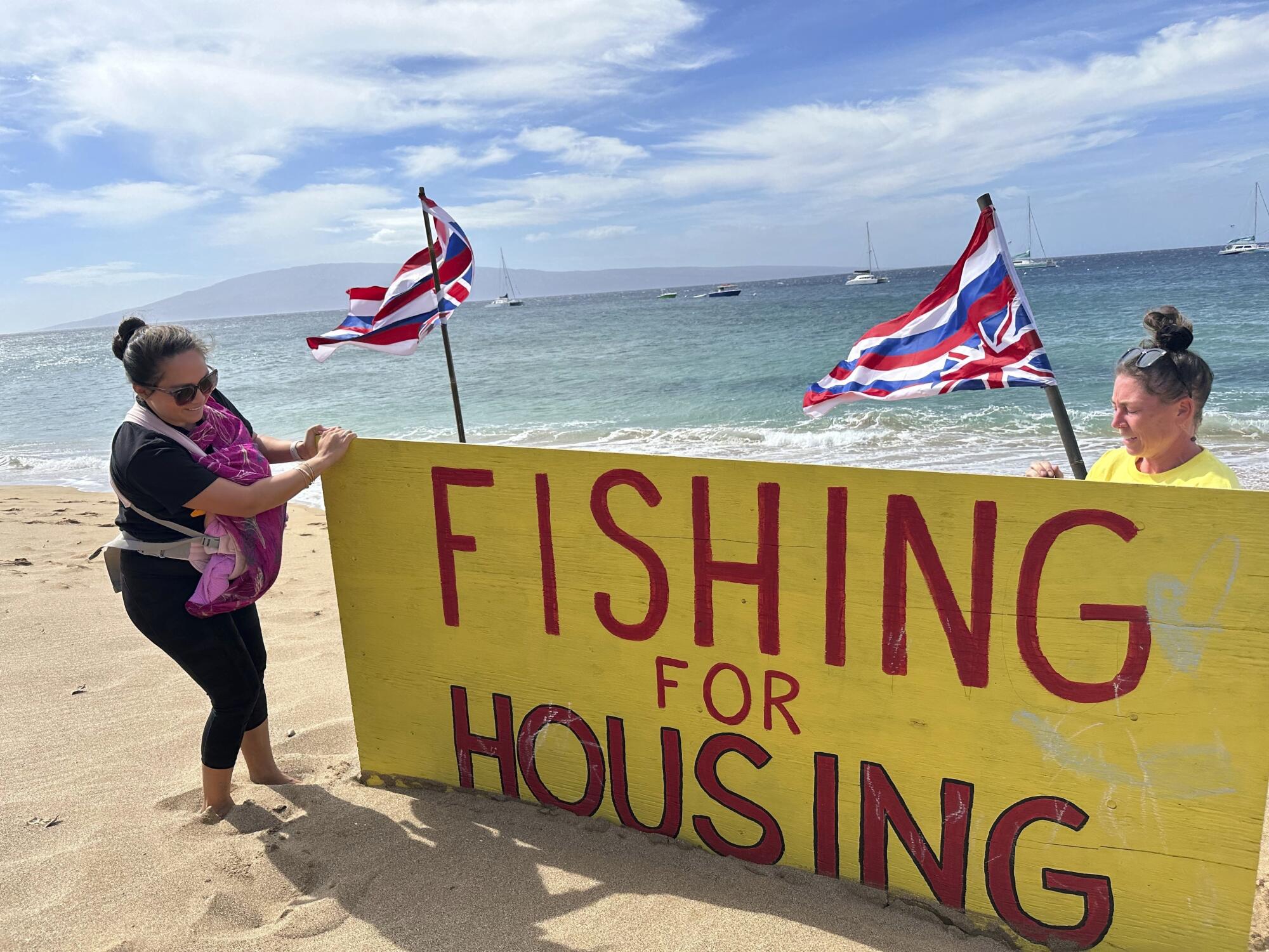 Two women adjust a large yellow homemade sign reading "Fishing for housing" on a beach as the wind blows 2 Hawaiian flags