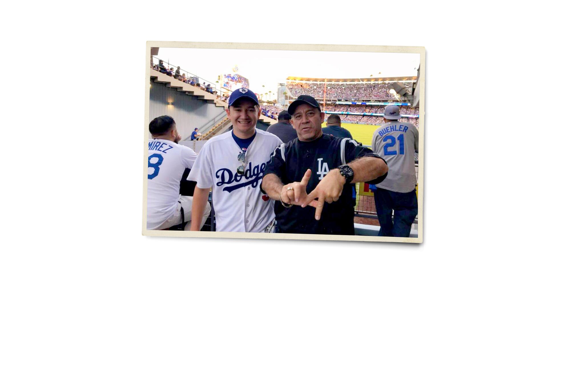 A photograph of a two men, one making the L.A. finger sign, both in Dodger gear at Dodger Stadium 