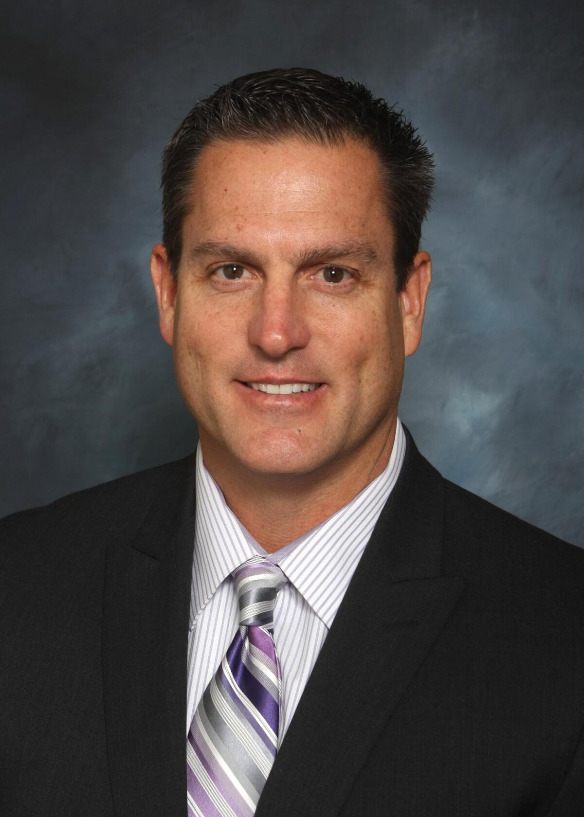 A head and shoulders portrait of a dark-haired man in a suit and tie, smiling and looking at the camera.