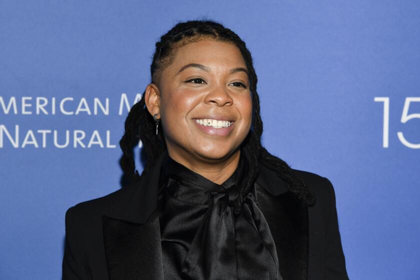 Punkie Johnson with braids wearing a black suit jacket and a black silk blouse. She poses in front of a blue backdrop