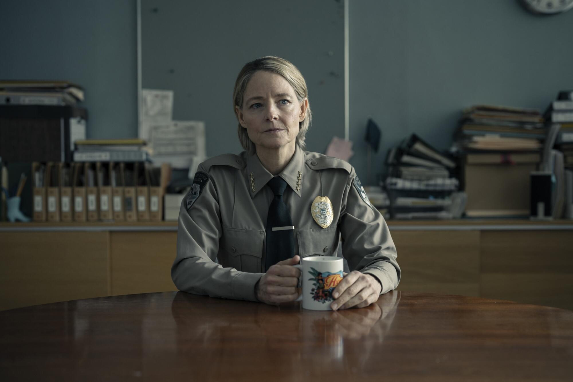 A woman in a police uniform holds a coffee mug while sitting at a table in "True Detective: Night Country." 