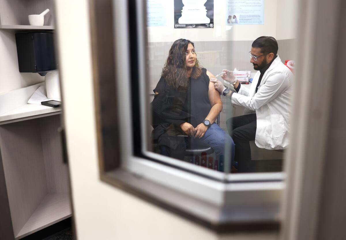 A man in a white coat gives woman seated in a chair a shot in the arm