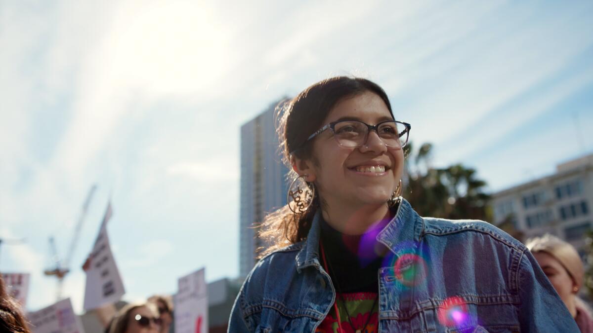 A smiling young woman with glasses stands outdoors with tall buildings and other women behind her