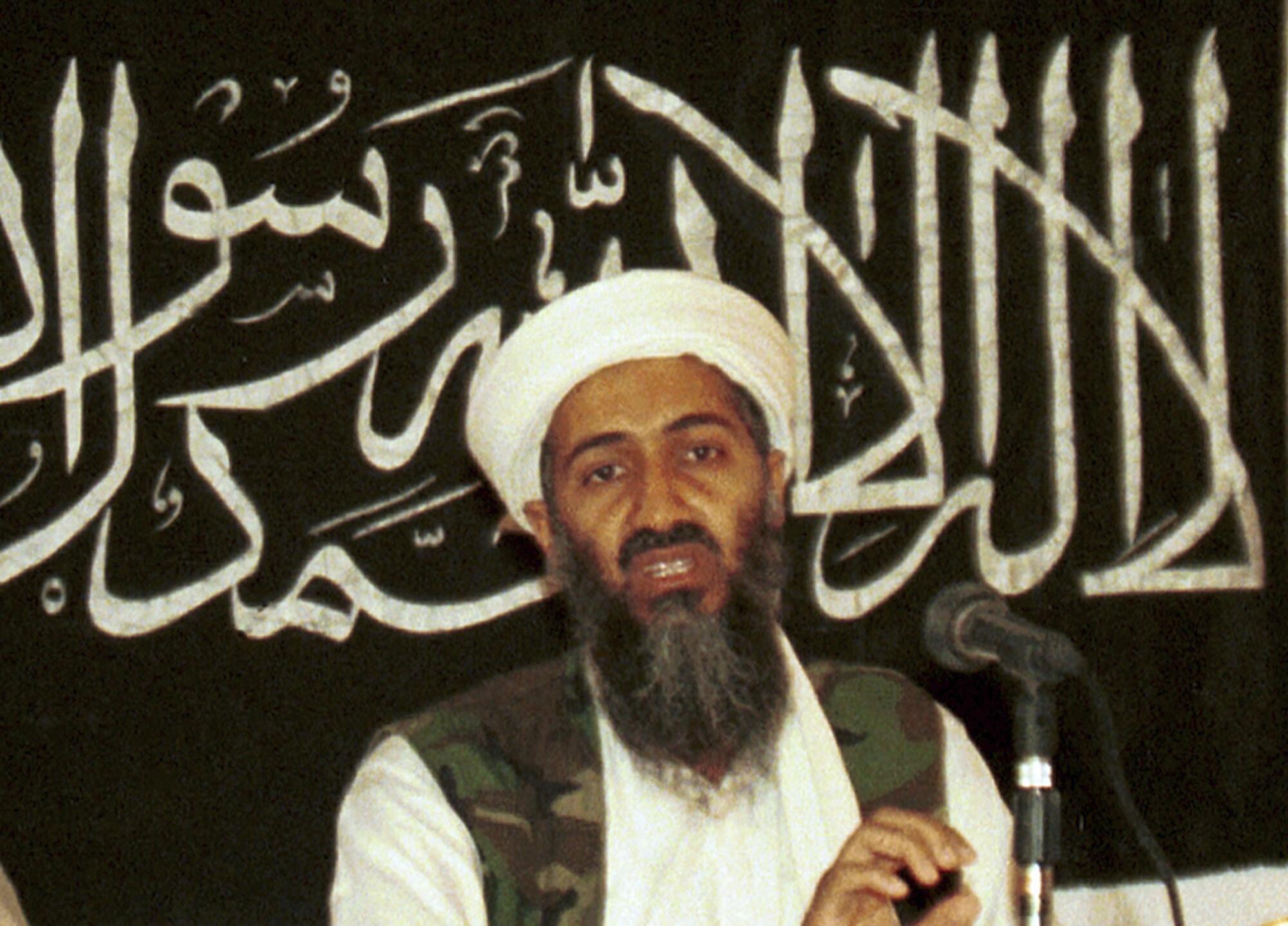 A bearded man in white turban and camouflage vest speaks before a microphone. Behind him is Arabic script on a dark backdrop.