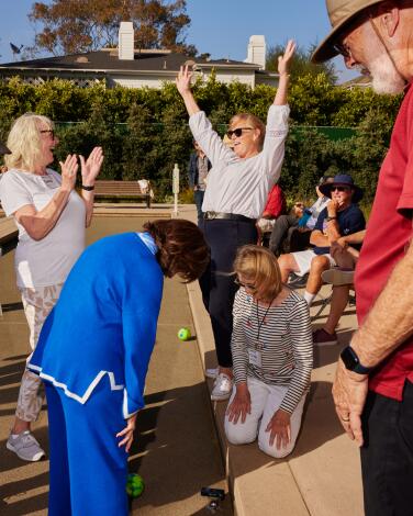 People kneel, stand and triumphantly raise their arms on an outdoor bocce court. 