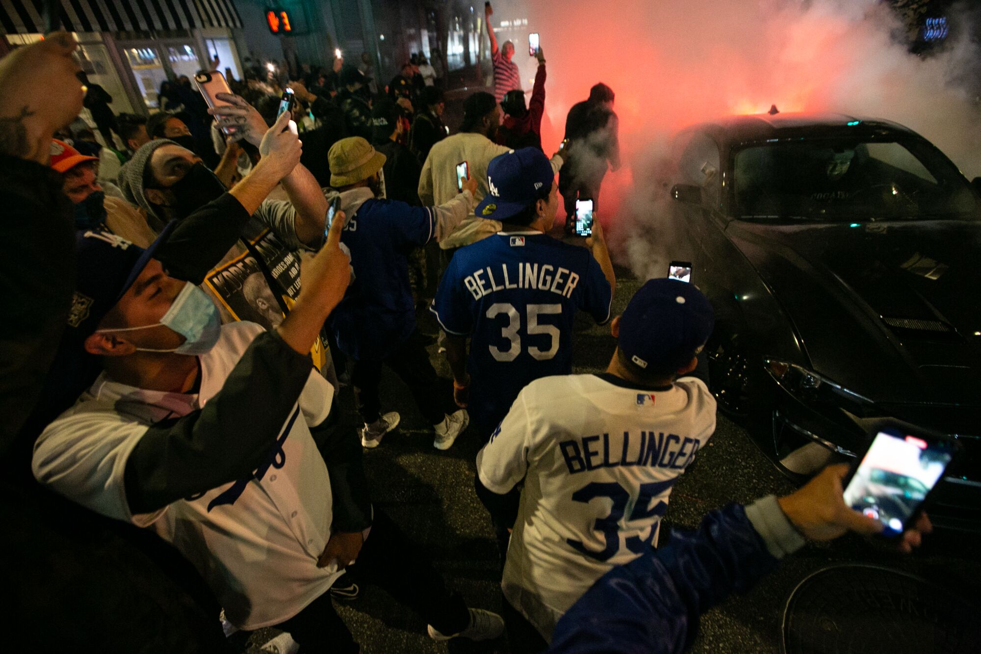 Fans in L.A., some in Dodgers jerseys, crowd together near a car, taking photos as smoke billows from its tires. 