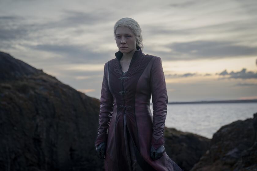 Rhaenyra Targaryen in a long coat standing on rocky terrain overlooking water