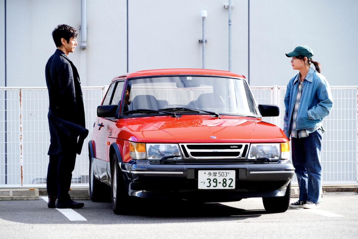 Two people stand on either side of a parked red compact car.