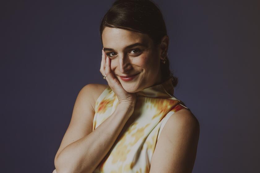 Actor Jenny Slate poses in a sleeveless yellow and white dress, one hand resting on her chin.