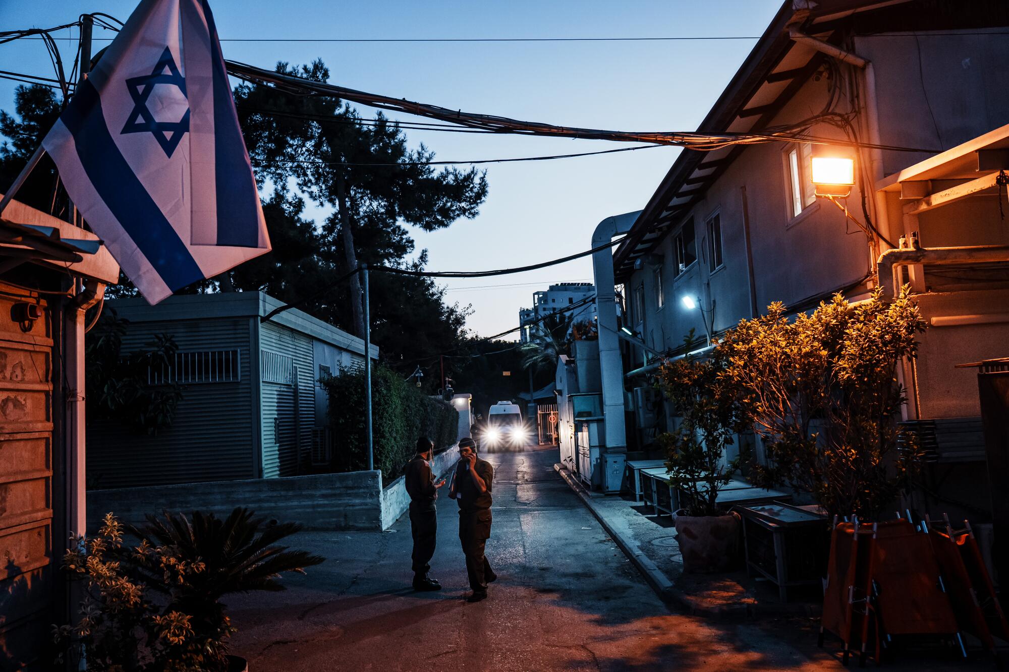 Two people stand on a path between buildings as a van with its lights on approach in the distance 