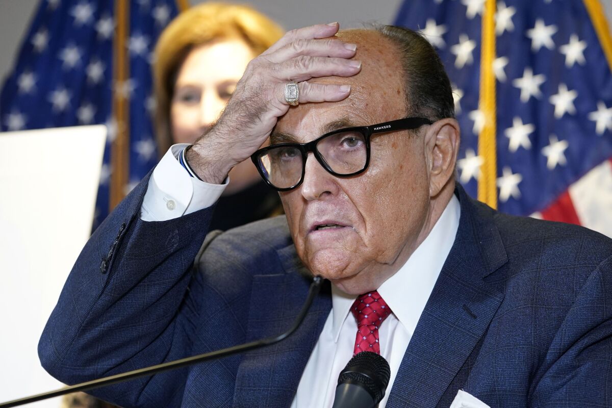 Rudy Giuliani, wearing a blue suit and red tie, presses his right hand to his forehead.