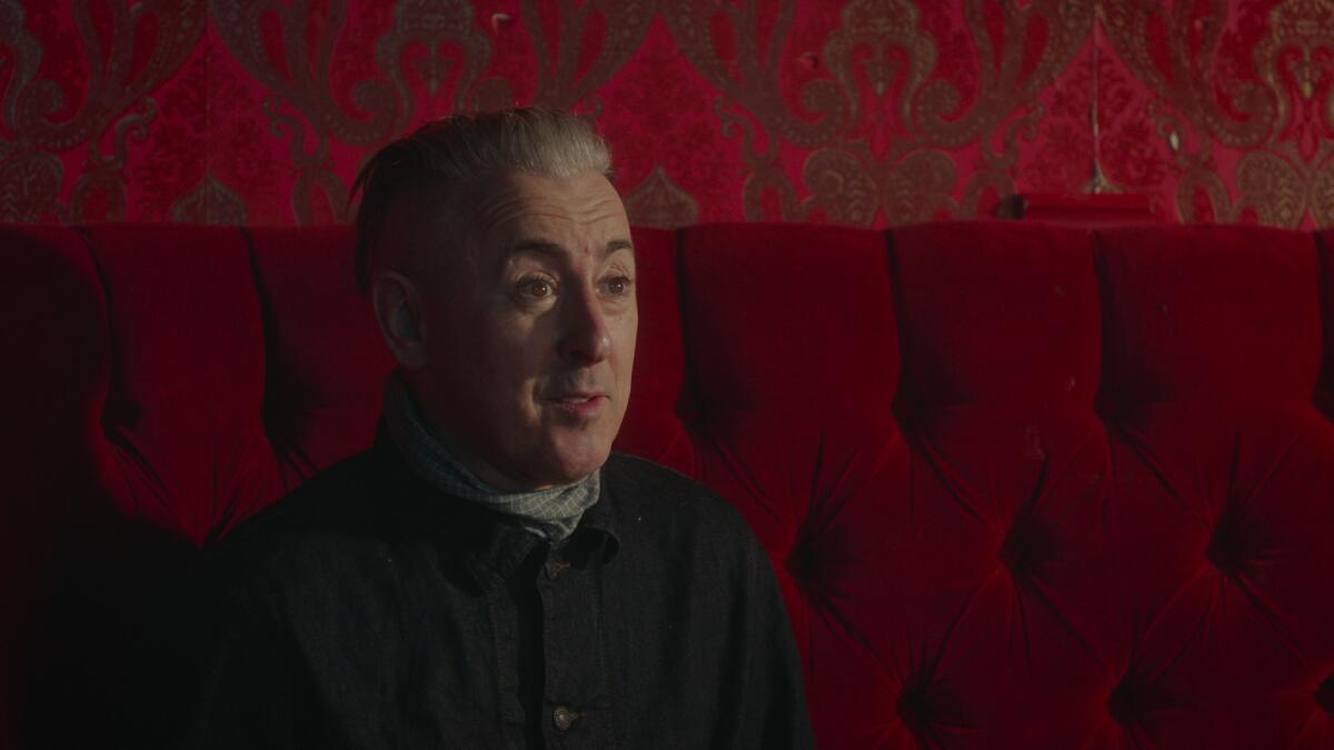 Alan Cumming in a dark shirt sitting on a red velvet sofa with red wallpaper behind him.