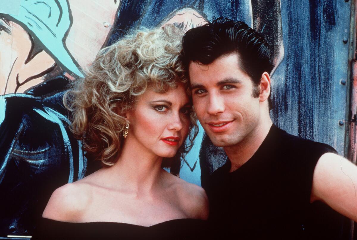 A blond woman with teased curly hair posing in a black bodysuit next to a man with quaffed brown hair posing in a black shirt