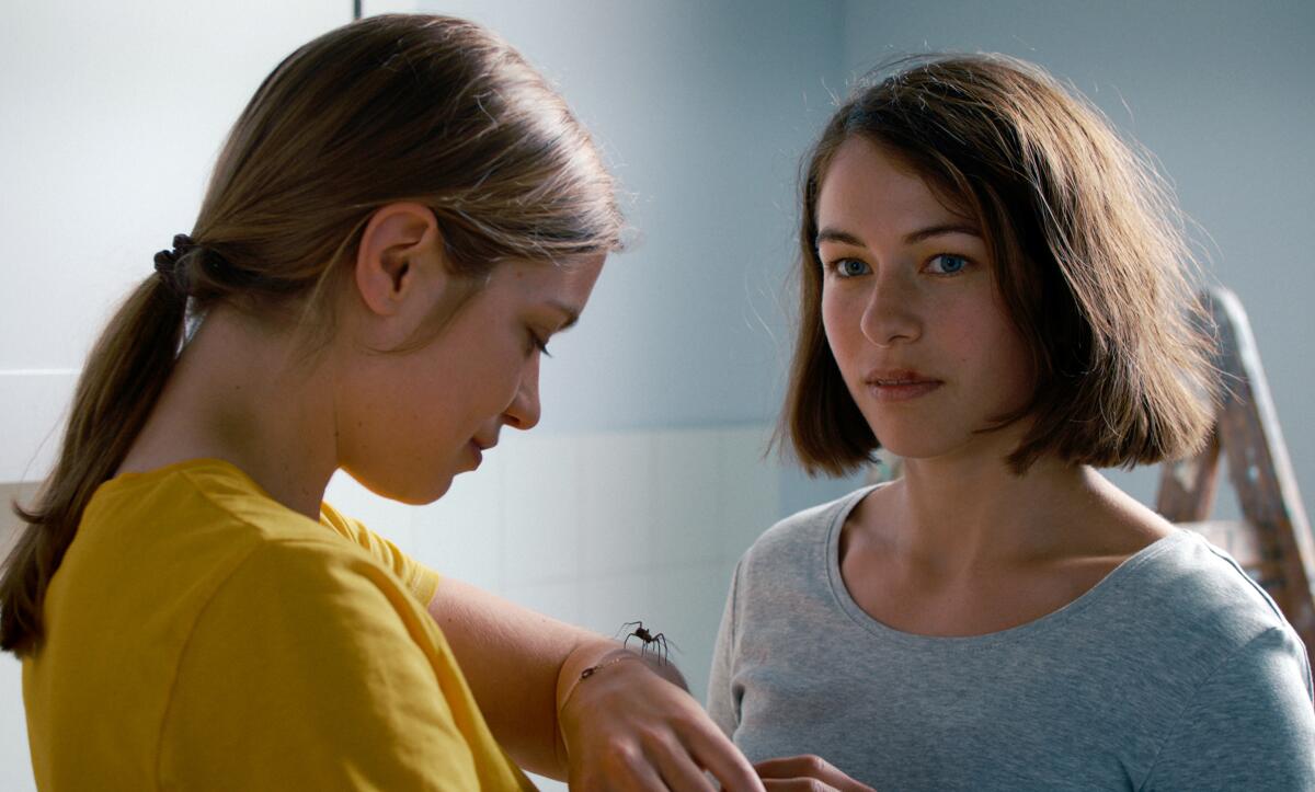 Two young women stand together; one looks at a large spider on her wrist.