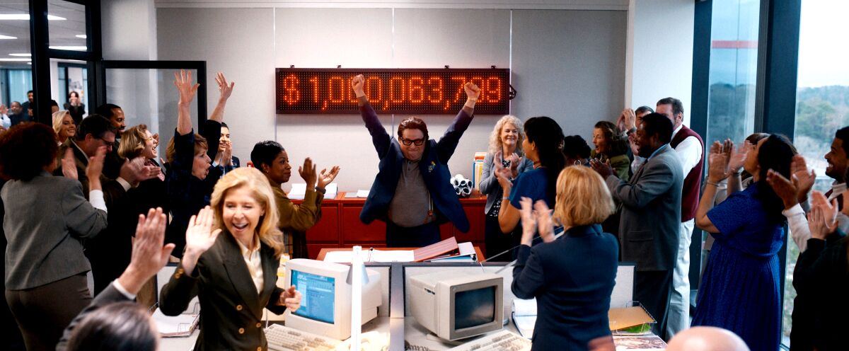 A man stands in an office, arms raised, as his employees cheer rising profits