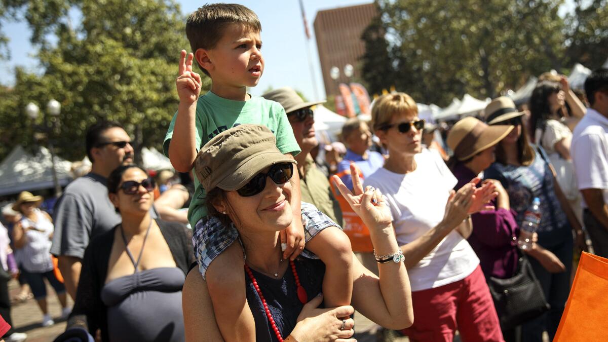 Among a crowd, a smiling woman in sunglasses has a small boy on her shoulders. They both make the peace sign.