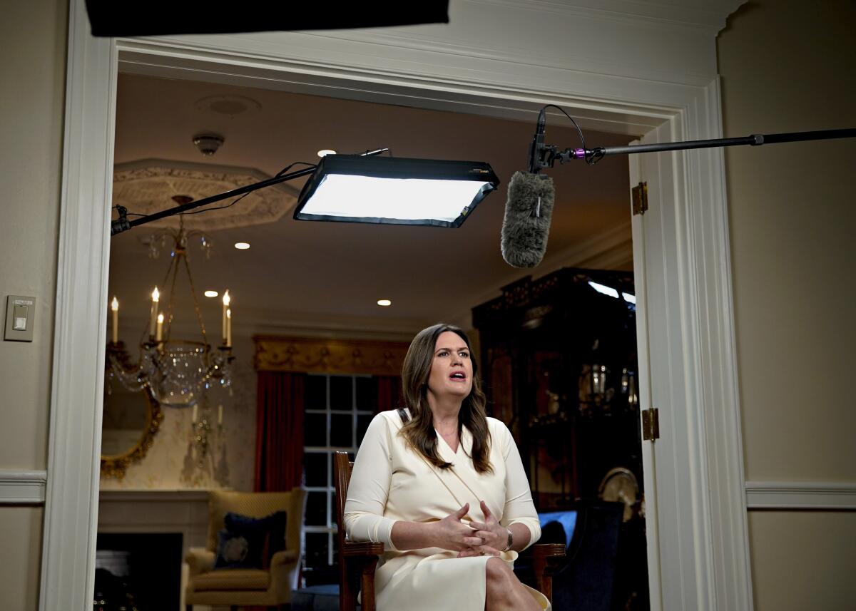 A woman with long brown hair sits in a chair, lit by TV light, and speaks.